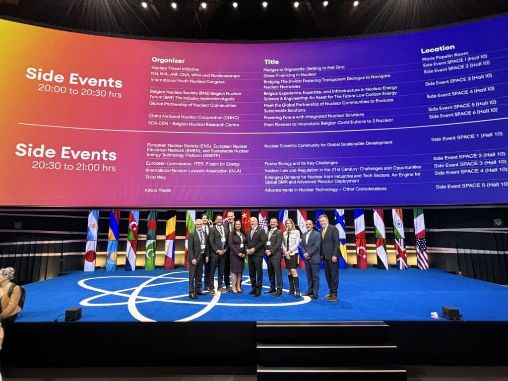 Group of professionals at the ARMA nuclear conference on stage with international flags and event schedule in the background.