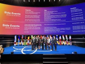 Group of professionals at the ARMA nuclear conference on stage with international flags and event schedule in the background.